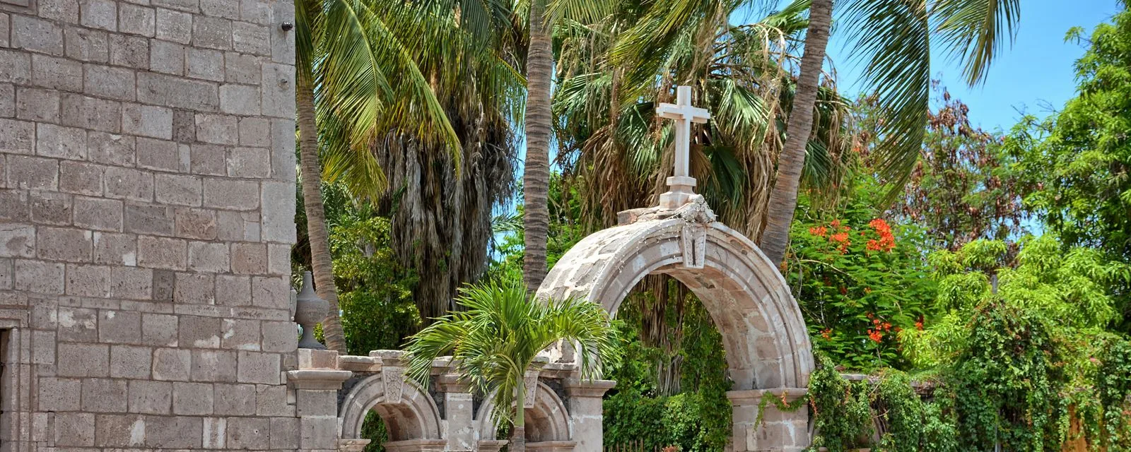 Arco de piedra rematado por una cruz en el conjunto de la misión de Nuestra Señora de Loreto, junto al muro de sillar y entre palmeras