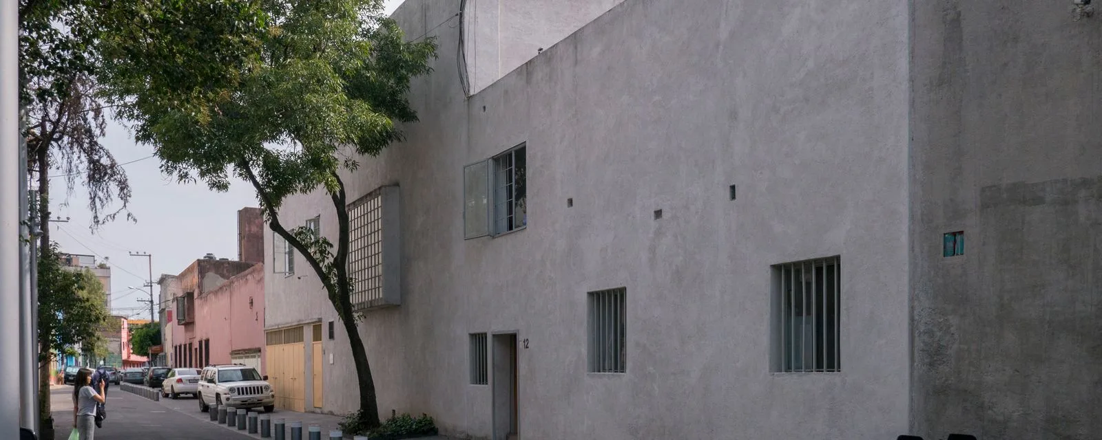 La fachada de la Casa Estudio Luis Barragán desde la calle: un muro liso de concreto sin ornamento, con ventanas altas enrejadas, entre casas del barrio