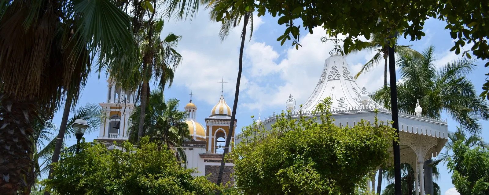 La plaza de Comala entre palmeras: el kiosco blanco de herrería a la derecha y las torres blancas con cúpula dorada de la parroquia al fondo