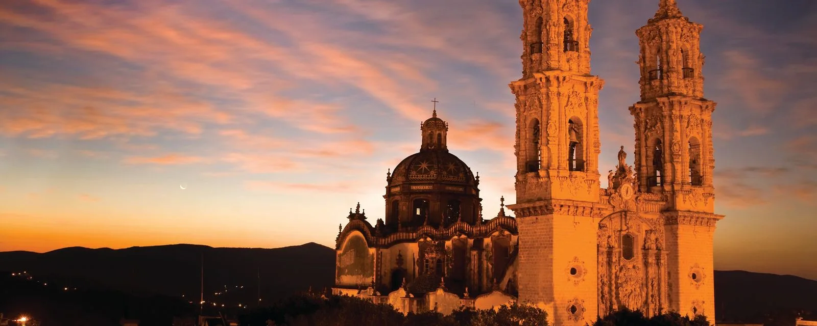 El templo de Santa Prisca de Taxco al atardecer: sus dos torres churriguerescas y su cúpula de azulejo iluminadas contra un cielo naranja