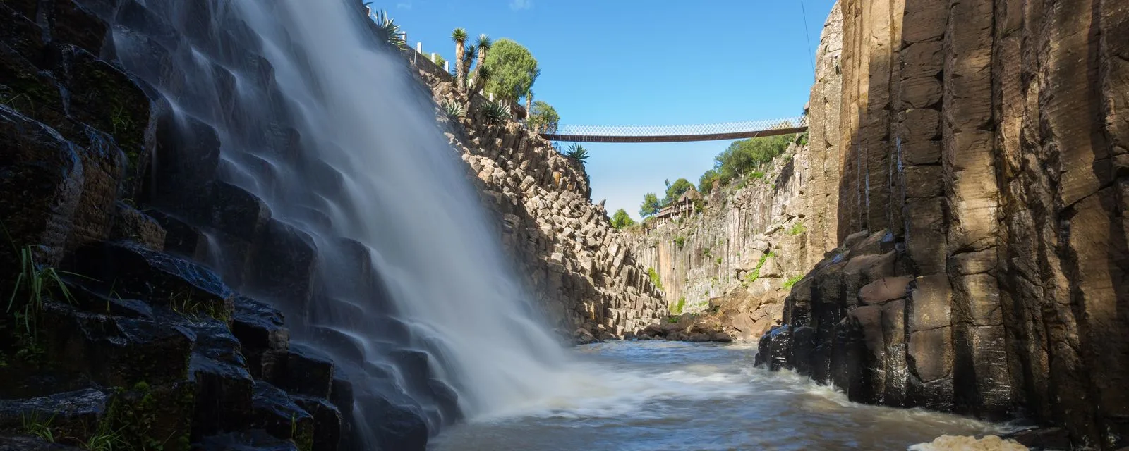 La barranca de los Prismas Basálticos desde abajo: la cascada cayendo a la izquierda, las columnas de basalto a la derecha y el puente colgante cruzando arriba