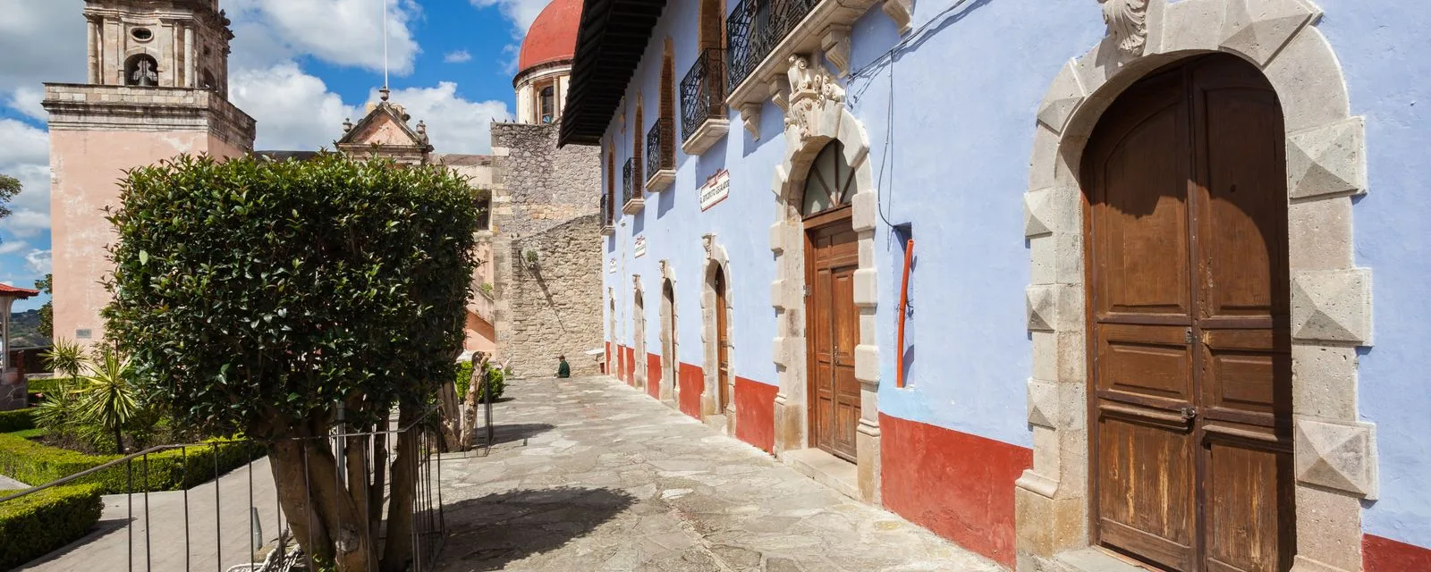 Una calle empedrada de Mineral del Chico entre una fachada azul con portones de madera y, al fondo, la parroquia rosa con su cúpula de teja