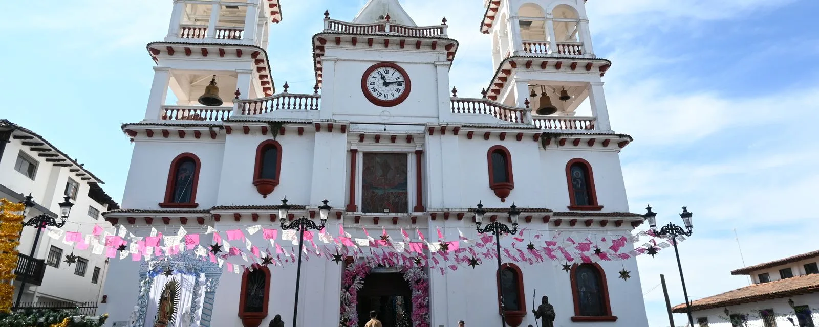 La parroquia de San Cristóbal de Mazamitla: fachada blanca con remates de teja roja y campanarios gemelos, con papel picado rosa cruzando el atrio