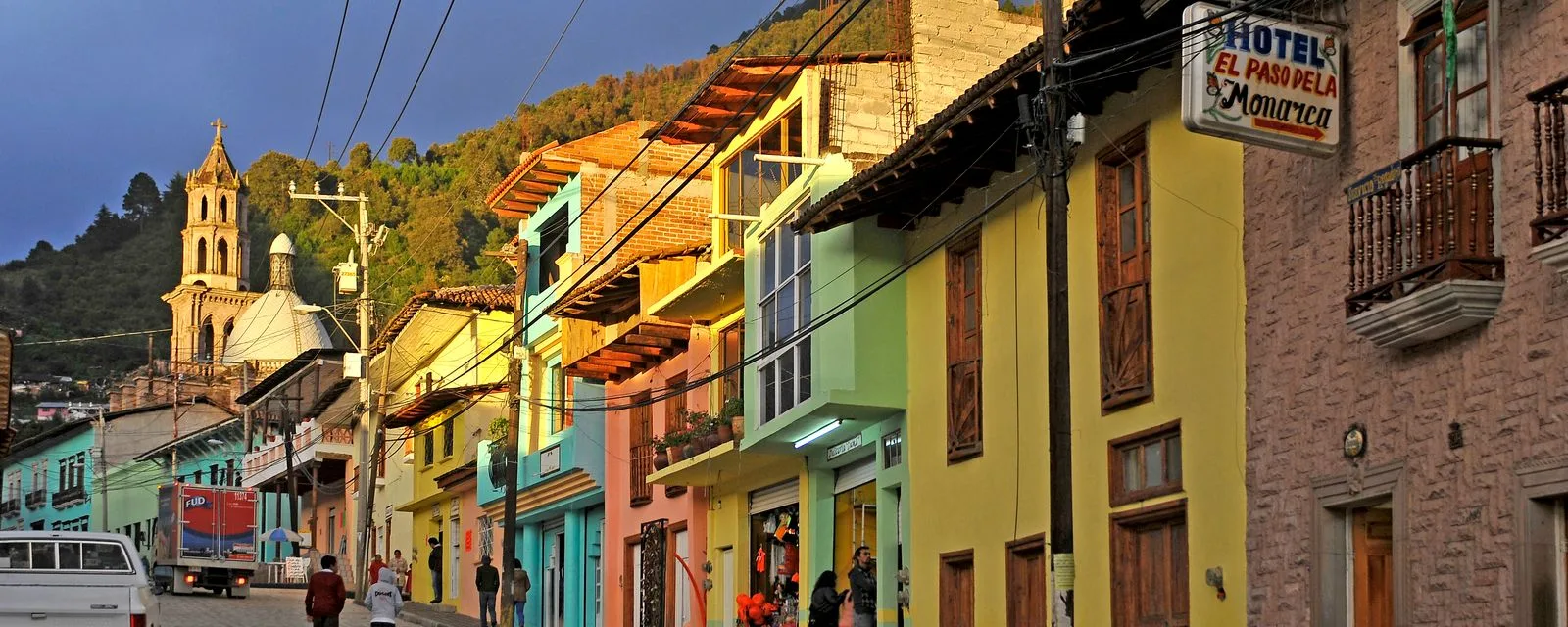 La calle principal de Angangueo al atardecer: casas de colores con balcones de madera subiendo la cuesta y la torre de la iglesia sobre el cerro al fondo