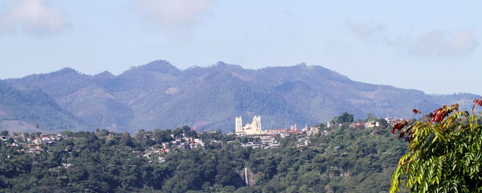 Xicotepec visto desde la ladera de enfrente: el caserío sobre el lomerío verde con la parroquia amarilla al centro y la sierra al fondo