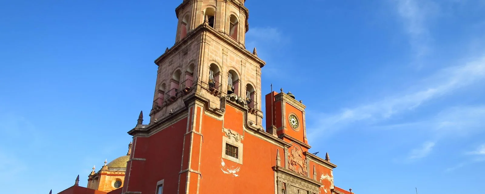 Templo del centro histórico de Querétaro visto desde la calle: torre de cantera, fachada de muro rojo con reloj y cúpula de azulejo contra el cielo despejado