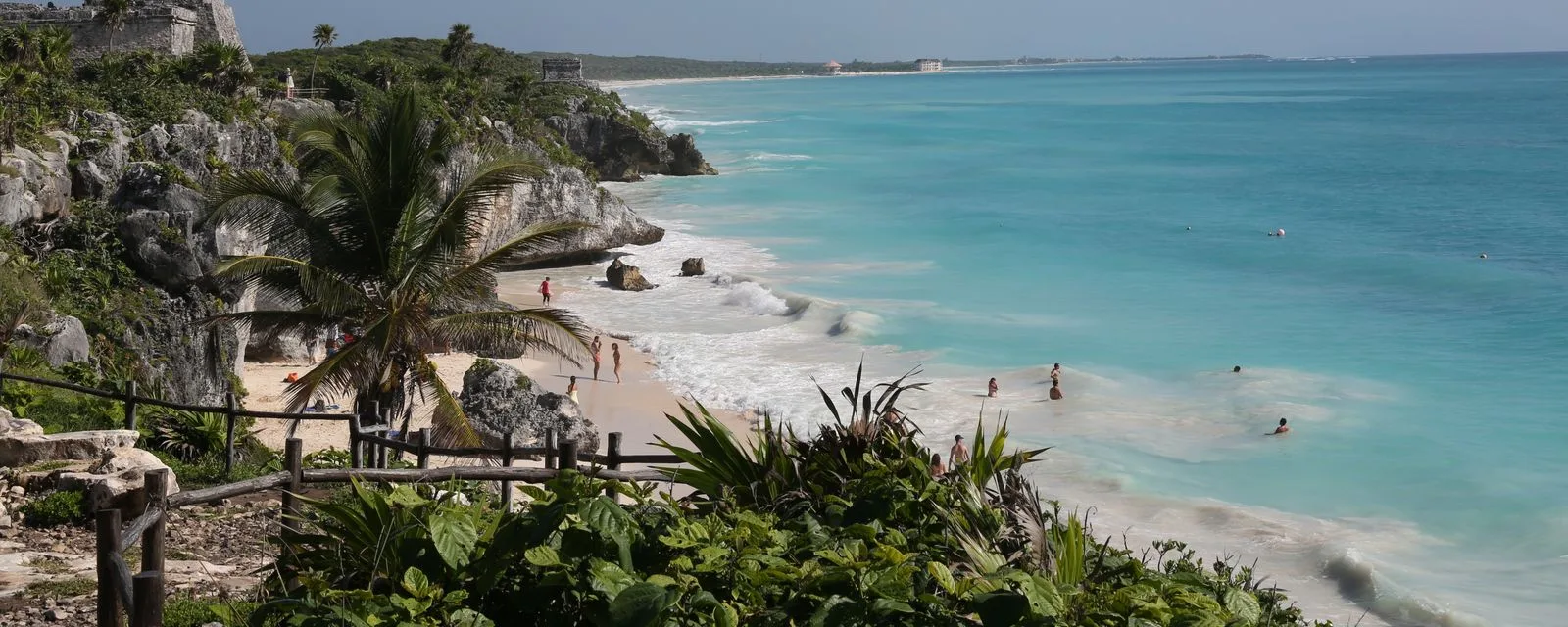 La playa al pie del acantilado de Tulum vista desde el sendero: arena blanca, mar turquesa y la vegetación de la zona arqueológica en primer plano