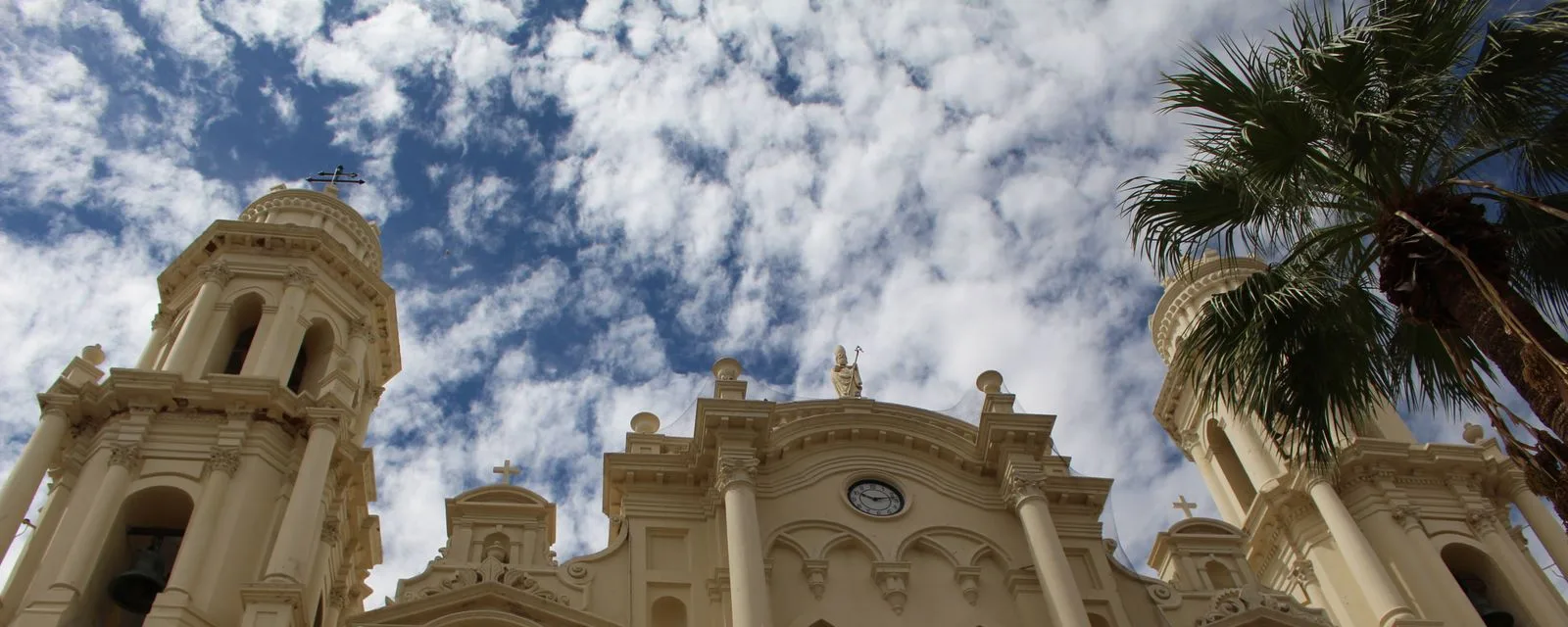 La Catedral de la Asunción de Hermosillo vista desde el pie de su fachada: torres, cúpula y frontón blancos recortados contra un cielo de nubes
