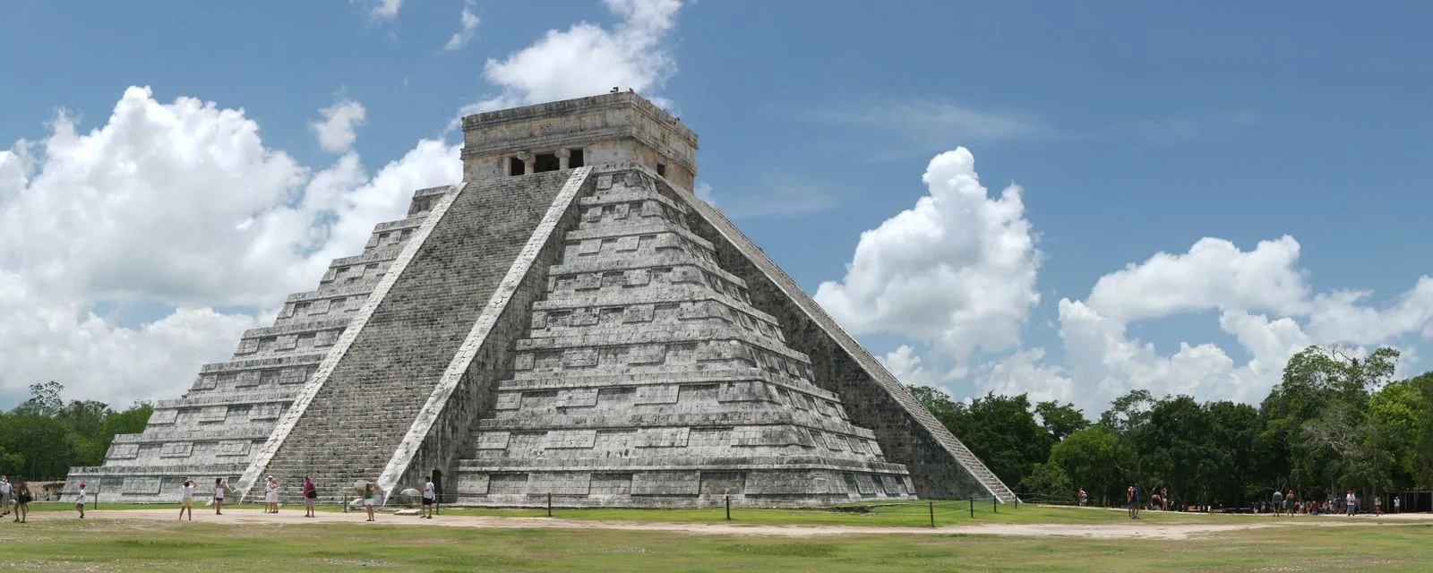 El Castillo de Chichén Itzá visto de esquina, con sus nueve cuerpos escalonados y dos de sus escalinatas, sobre la explanada de pasto